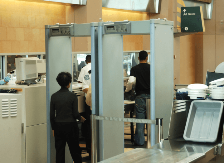 A woman in a grey polar neck jumper and black trousers walks through a metal detector at airport security. There are trays for passenger belongings to the right of the metal detector and to the left of it, there are airport security agents wearing blue rubber gloves. Beyond the metal detector, there is a sign on a brown wall that directors passengers to all departure gates.