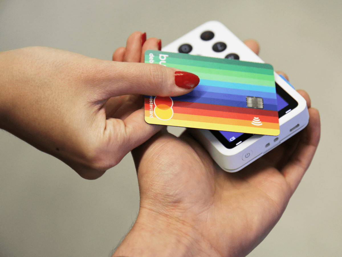 A hand with red nail polish holding a rainbow-designed Bunq card taps the card against a Contactless reader that is held by another person.