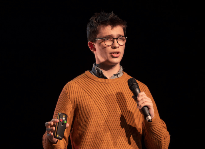 A young man in an orange jumper and thick-rimmed glasses stands on a stage, speaking into a microphone.