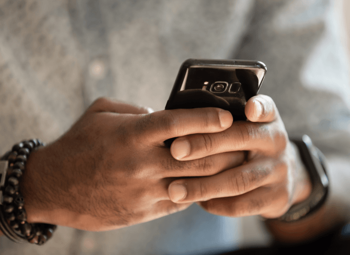 A man holding a mobile phone. His head is cropped from the image but he is wearing a grey sweater and a black watch on his left arm. On his right arm, he is wearing a beaded bracelet.