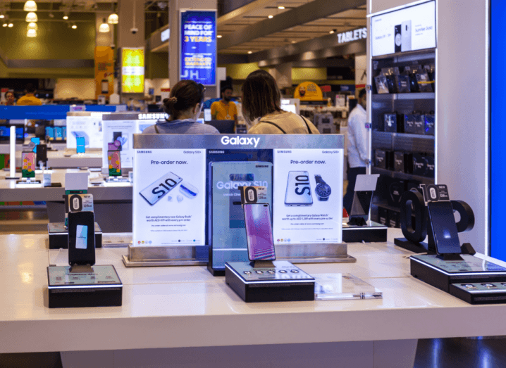 Different models of the Samsung Galaxy S10 on display at an event or convention. People are walking around in the background beyond the phones.