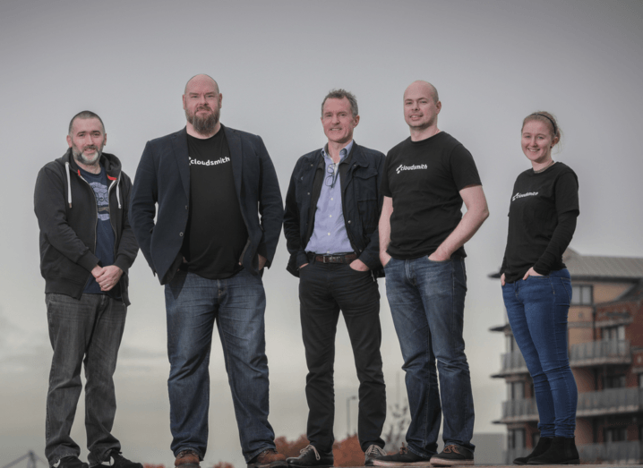 A group of five Cloudsmith employees stand in front of a grey sky in Belfast, all wearing black T-shirts with the white Cloudsmith logo on the front.