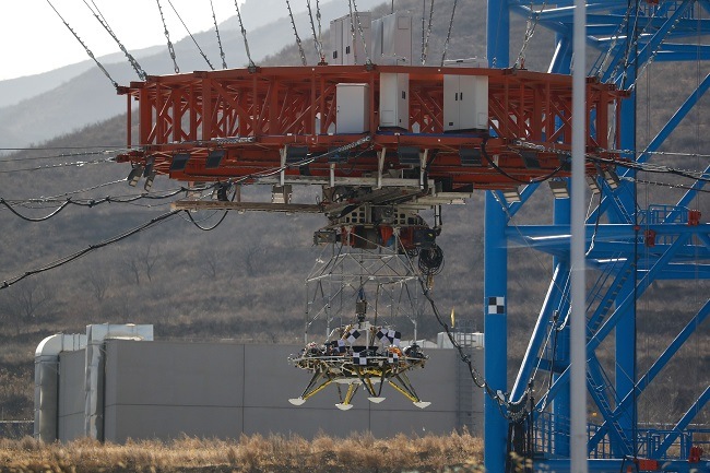 The lander suspended in mid-air by a red lifer beside a blue metal structure.