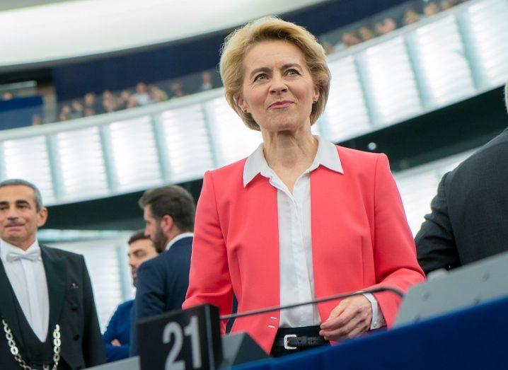 Ursula von der Leyen in a pink blazer smiling in the EU Parliament chamber.