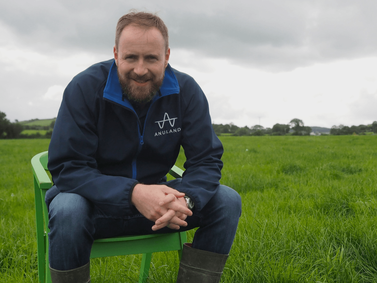 A bearded man with brown hair wearing a navy fleece and blue jeans with wellies sits on a green plastic charge in a large field full of grass.