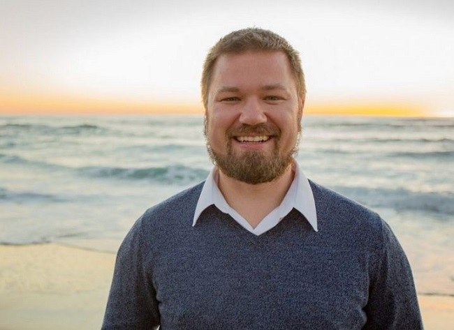 Erik Trask in a blue jumper and white shirt smiling against a sunset background by the sea.