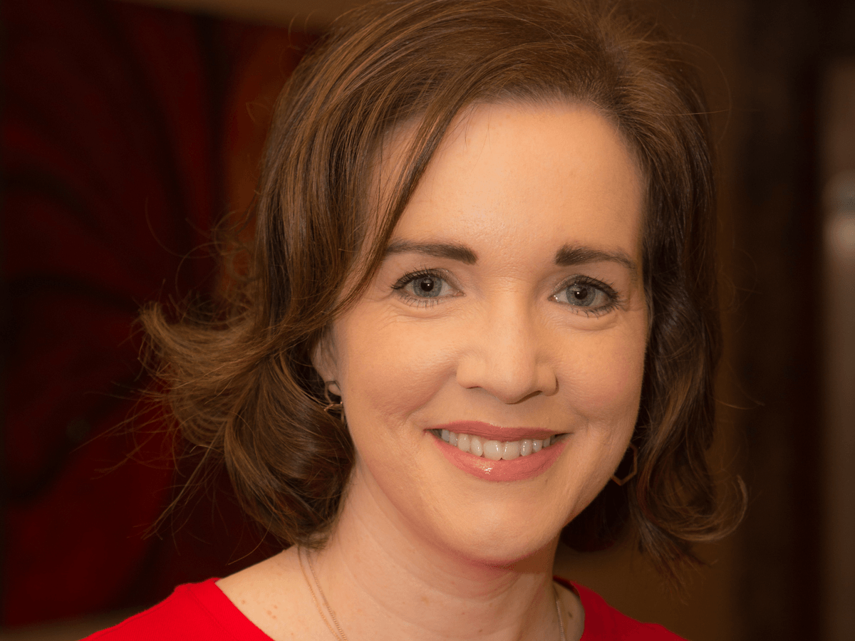 A woman with short, brown, curly hair wearing a red top smiles into the camera. 