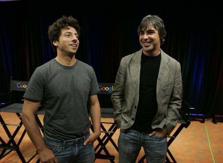 Sergey Brin and Larry Page standing beside each other smiling with fold-out chairs and a black curtain behind them.