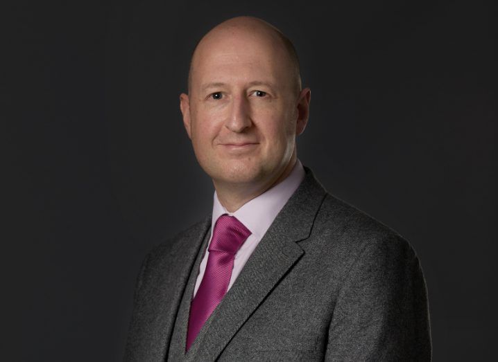 View of bald man in business attire, grey blazer, white shirt, purple tie, standing against dark background.