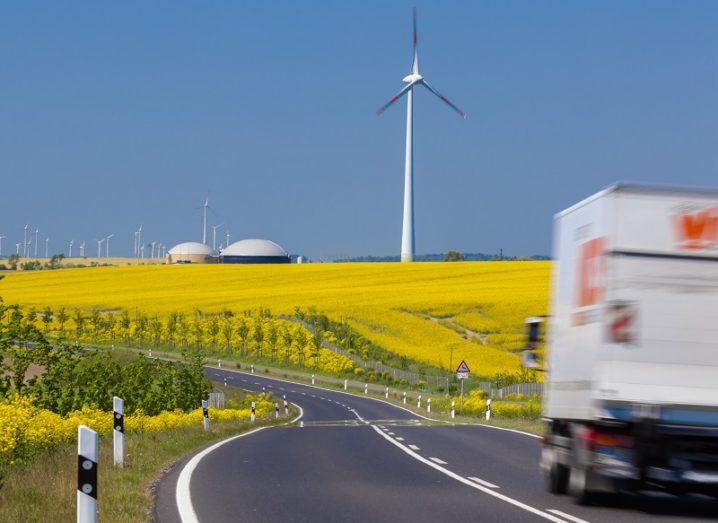 Blurred image of a passing truck on a empty road against a backdrop of yellow field and wind turbines.