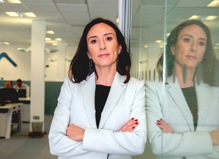 A confident woman in a white blazer leans against a glass wall in an office with her arms folded, her reflection clearly visible.