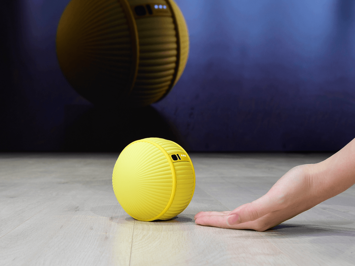 A yellow, tennis ball-sized robot rolls on the floor beside a human's hand. It has a small black camera on it.