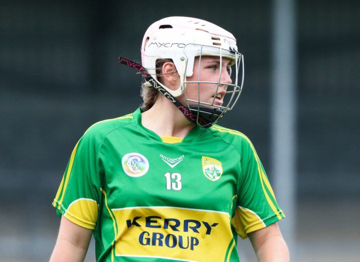 A Kerry camogie player in a green and gold jersey and protective helmet glances across the playing field.