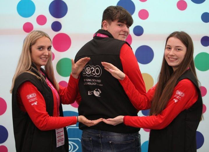 Three BT Young Scientist volunteers, with two pointing to the logo on the middle person's back.