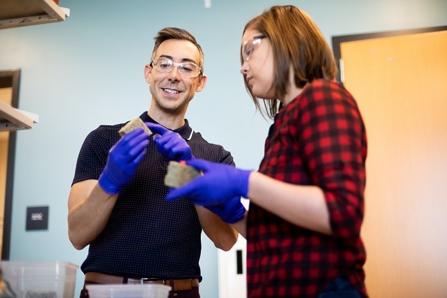 Wil Srubar and a lab student wearing purple gloves and safety goggles while holding the biomaterial in a lab. 