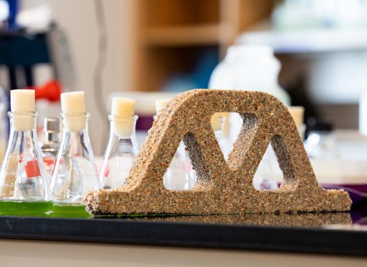 The bio-concrete moulded into a lattice brick shape on a lab table beside beakers filled with green liquid.