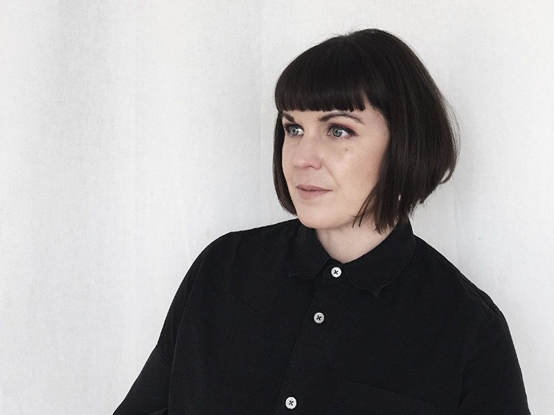 Woman with dark hair and a black shirt sits against a white wall.