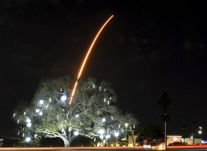 A tree by a roadside is lit up against a dark sky, and a bright red trail of the Falcon 9 rocket is seen emerging from behind it, rising into the stratosphere.