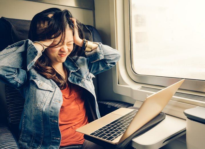 Woman in a denim jacket and orange t-shirt holding her head in her hands in frustration, while using a laptop on a train.