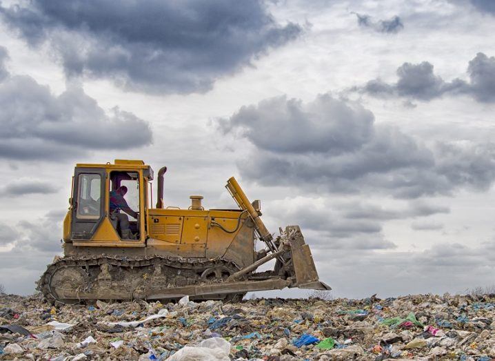 Ground shot of a bulldozer driving across a field of plastic waste against a grey sky background.