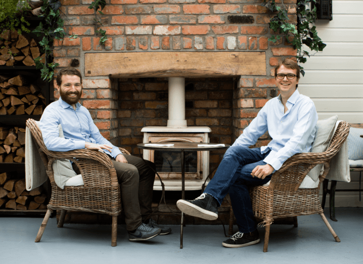 Two men in light blue shirts sit on wicker chairs in front of a stone fireplace.