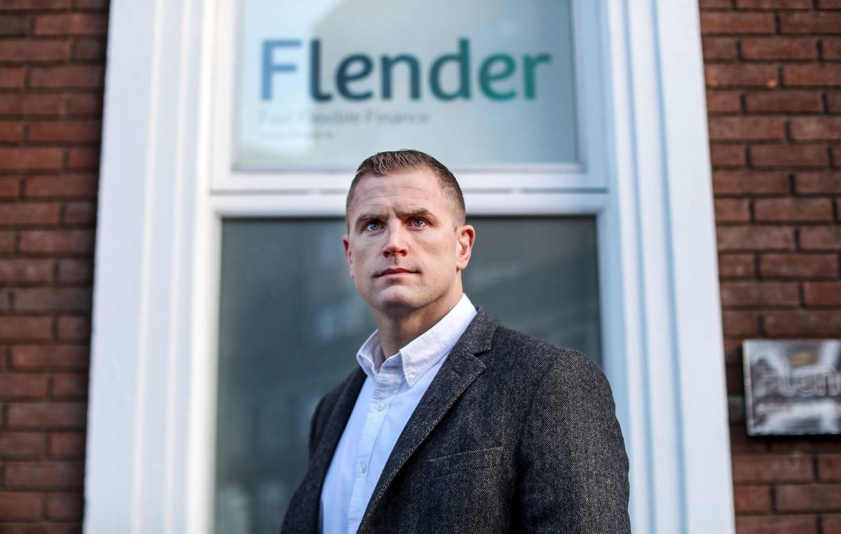 A man in a grey suit stands outside of a red brick building in front of the Flender logo.