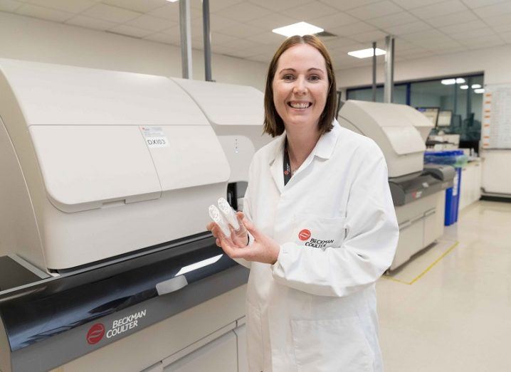 Joan Russell in a white lab coat smiling in front of manufacturing equipment at Beckman Coulter.