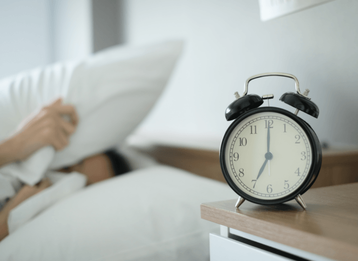 A person struggling to wake up, lying under a duvet with a pillow over their head, and an alarm clock on the bedside table.