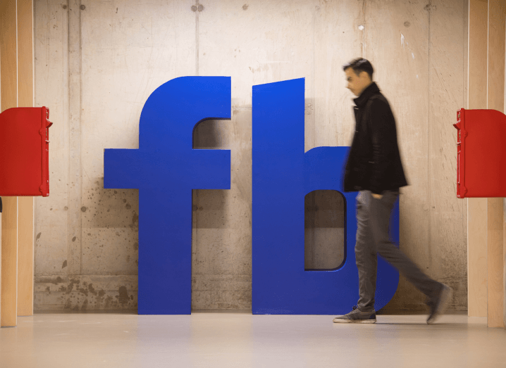 The Facebook logo on a wall in a grey concrete room where a man is walking past wearing a black jumper and blue jeans.