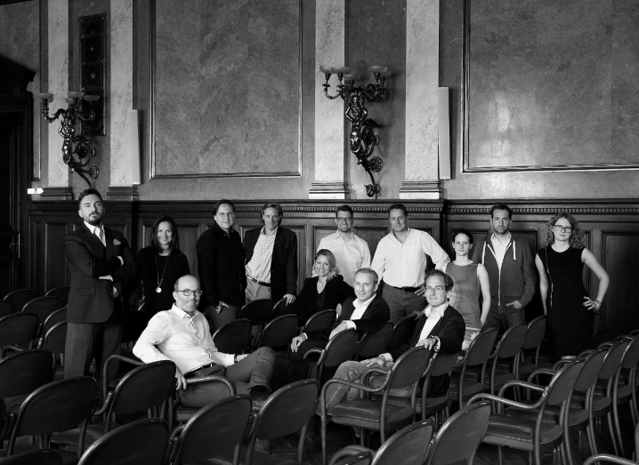 A large group of men and women stand in a room together in front of rows of chairs. Some of the people are sitting on the chairs. The photograph is in black and white.