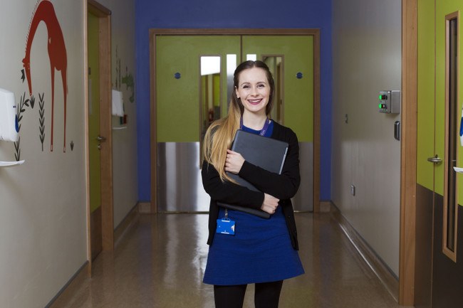 A young woman stands in a hospital corridor smiling and holding a closed laptop.