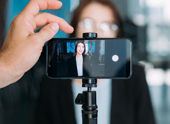 Woman wearing a black blazer and glasses standing in front of a smartphone camera with another hand about to press record.