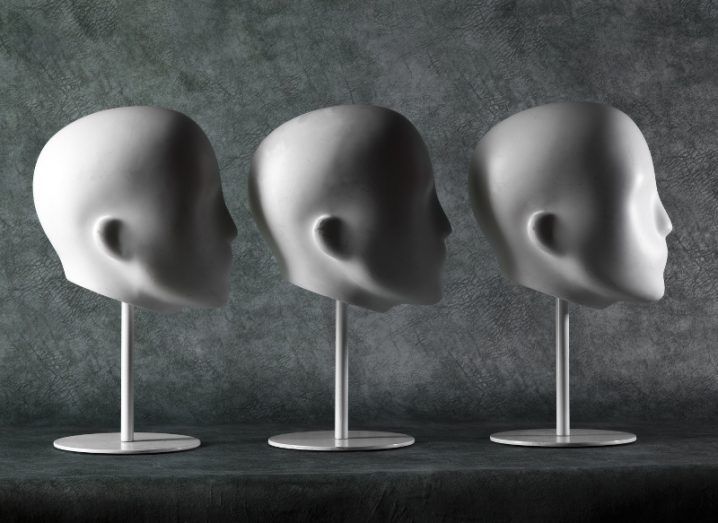 A row of three white plaster mannequin heads, all facing to the right against a grey-green stone background.