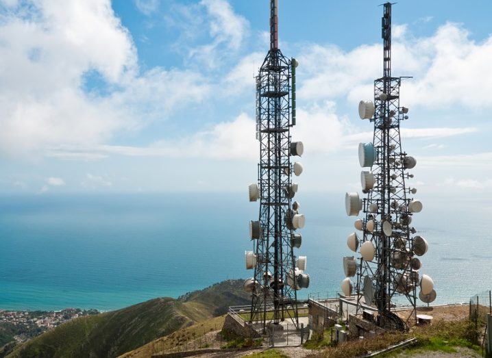 Two mobile network towers on top of a hill overlooking an ocean.