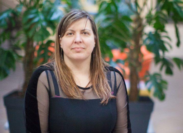 A woman with long hair and a black top sits in a bright room with potted plants behind her.