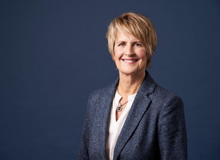 A woman with blonde hair and a blue suit stands against a blue background.