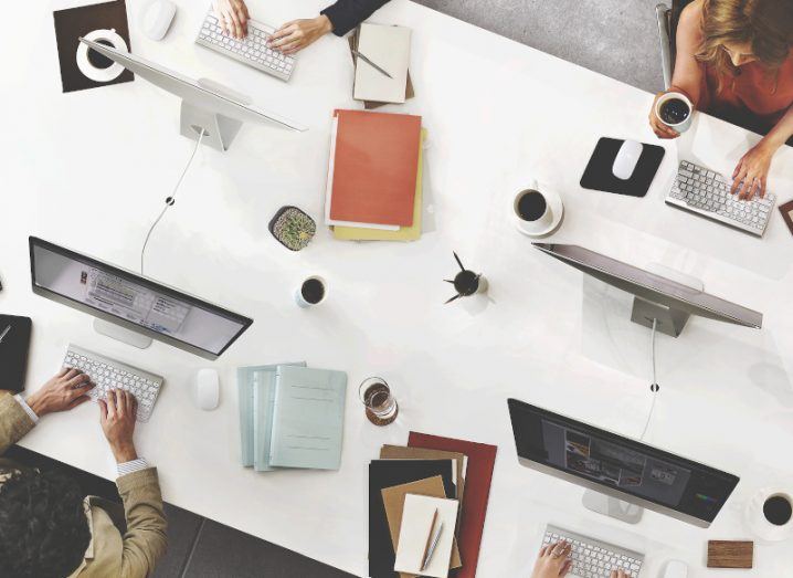 A bird’s eye view of a white table with four people working on computers. There are also notebooks and coffee on the table.