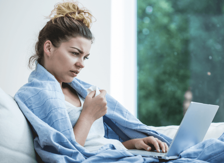 A sick woman using a laptop while wrapped in a blue blanket.