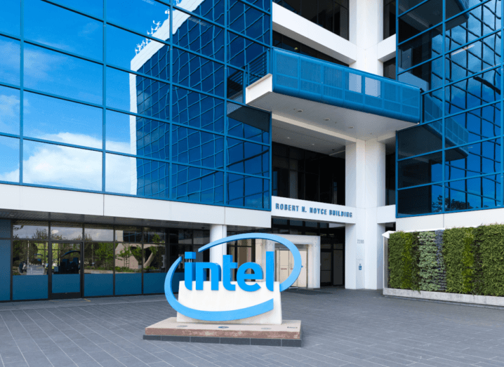 The Intel logo displayed out of a large, glass-fronted office building in Santa Clara, California. To the right of the sign there are hedges. From the reflection in the building, a sunny, blue sky is visible.