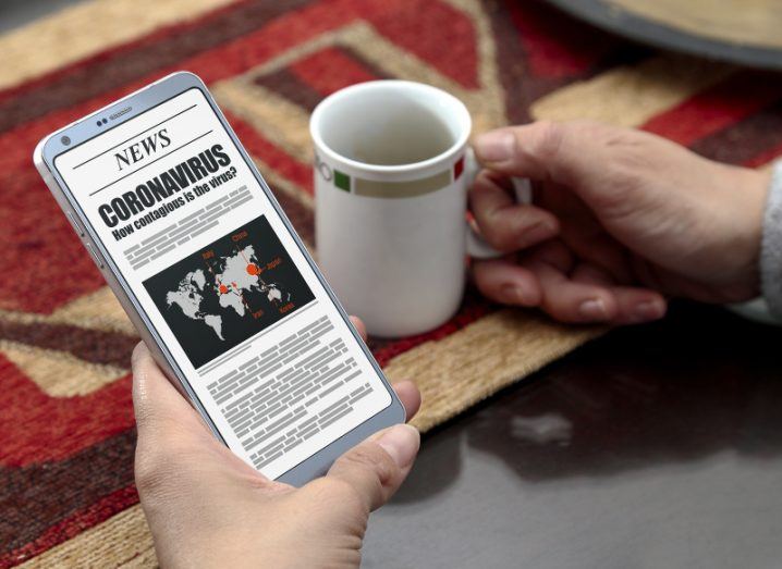 A woman holds a smartphone in one hand to read a news story about coronavirus while drinking a cup of coffee.