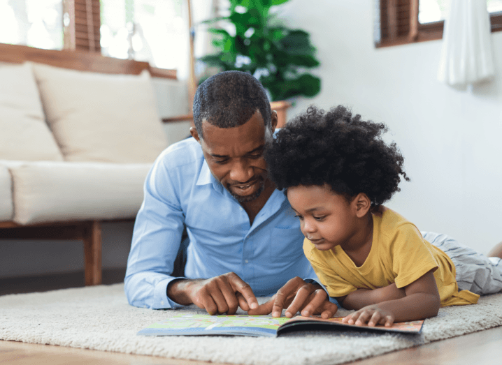 A man in a blue shirt lying down on the floor in a sitting room, helping a child in a yellow t-shirt to read.