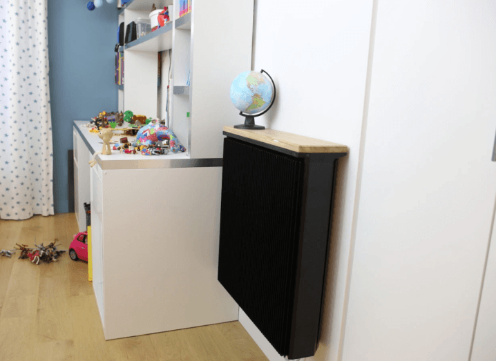 A black radiator mounted to a wall in a child's bedroom beside a desk. There are toys all over the desk and floor.