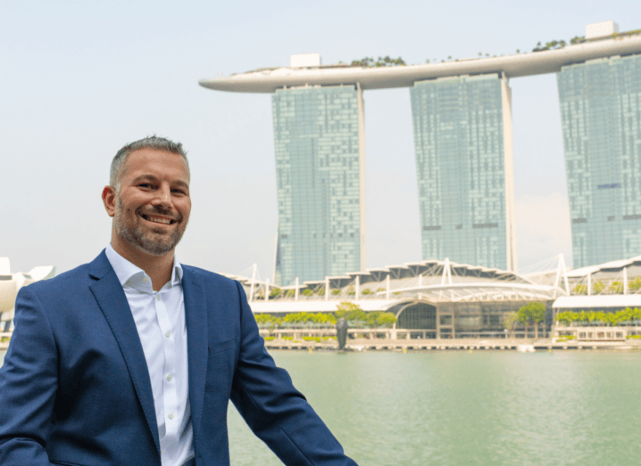 A man wearing a navy suit smiling in front of the famous Marina Bay Sands building in Singapore.