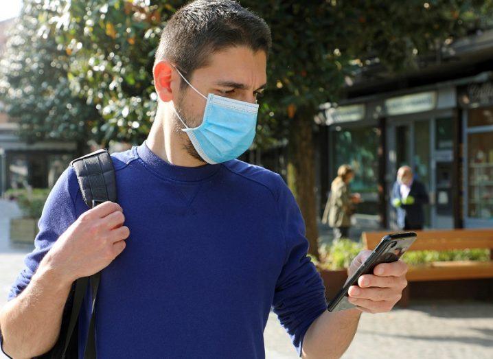 Man in a blue jumper wearing a face mask looking at his phone in a public place.