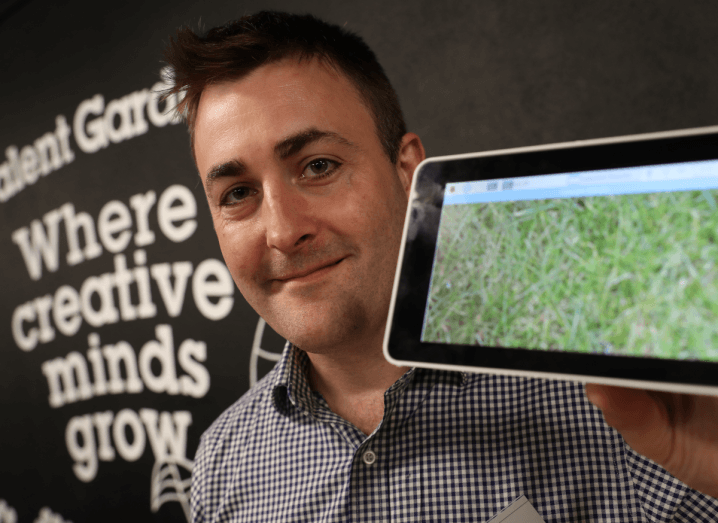 A man with brown hair holding a tablet and standing in front of a wall with the Talent Garden logo on it.