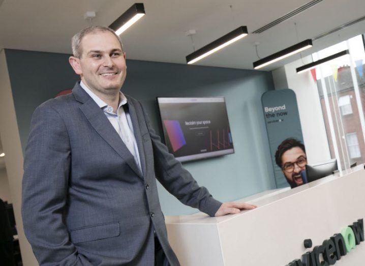 Mark Cockerill stands in a suit at a reception desk that says 'ServiceNow' on the front of it.