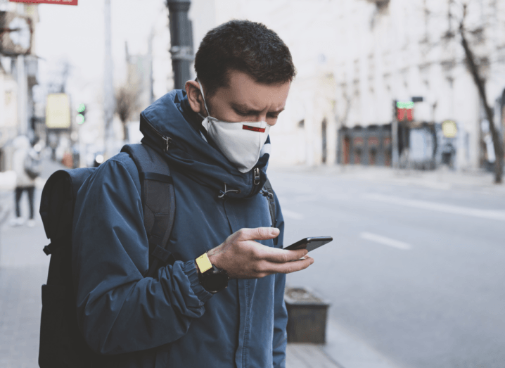 A young man wearing a white mask with a blue jacket stands on a street looking at his phone. He has brown hair and is also wearing a black backpack.