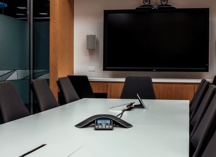 A conference room with a television mounted on the wall and a phone in the centre of a table, surrounded by seven chairs.