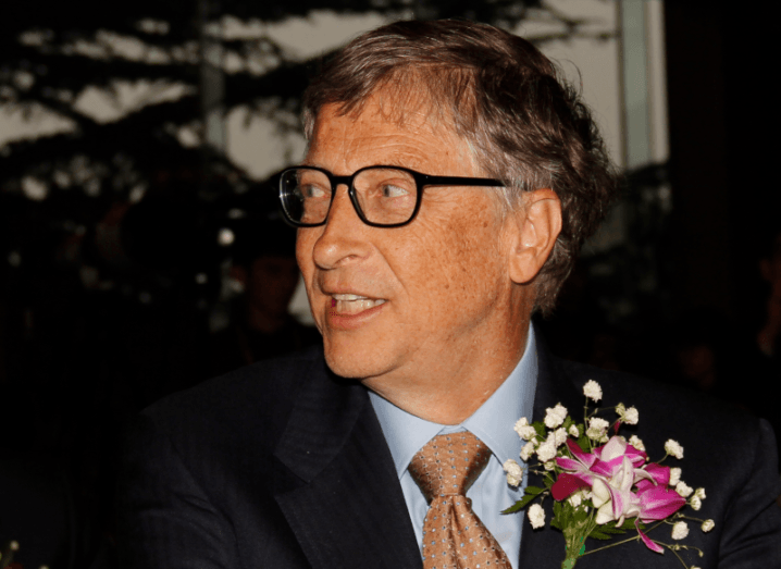 Bill Gates wearing black glasses and a suit with flowers attached to his lapel.