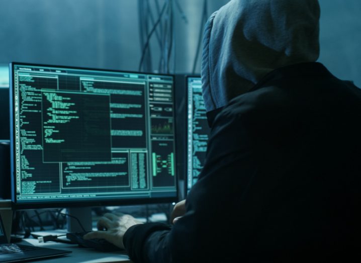 A hooded hacker sits at desk in a dark room. He is working at a monitor with a lot of green code on it.